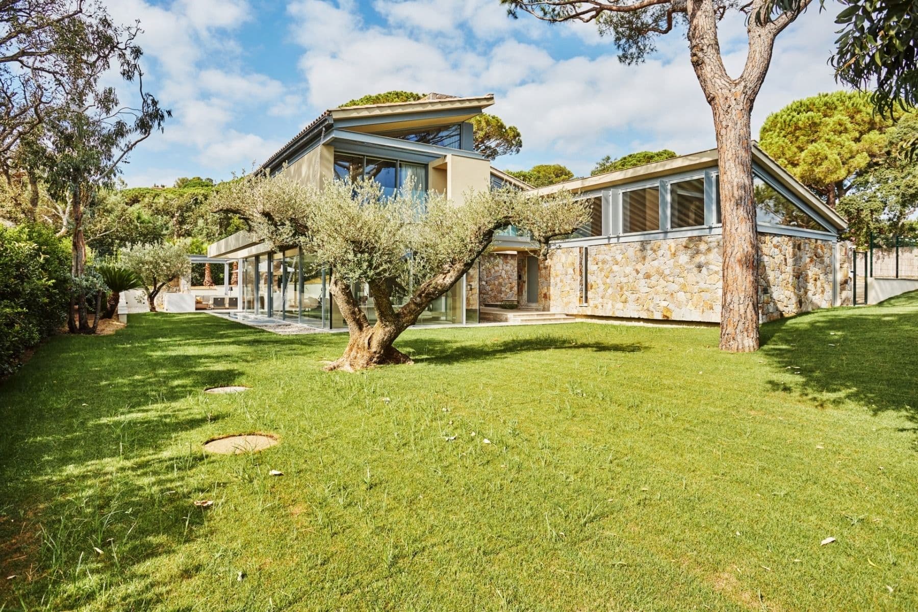 Modern stone and glass villa with pitched roofs set among pine trees and a manicured lawn