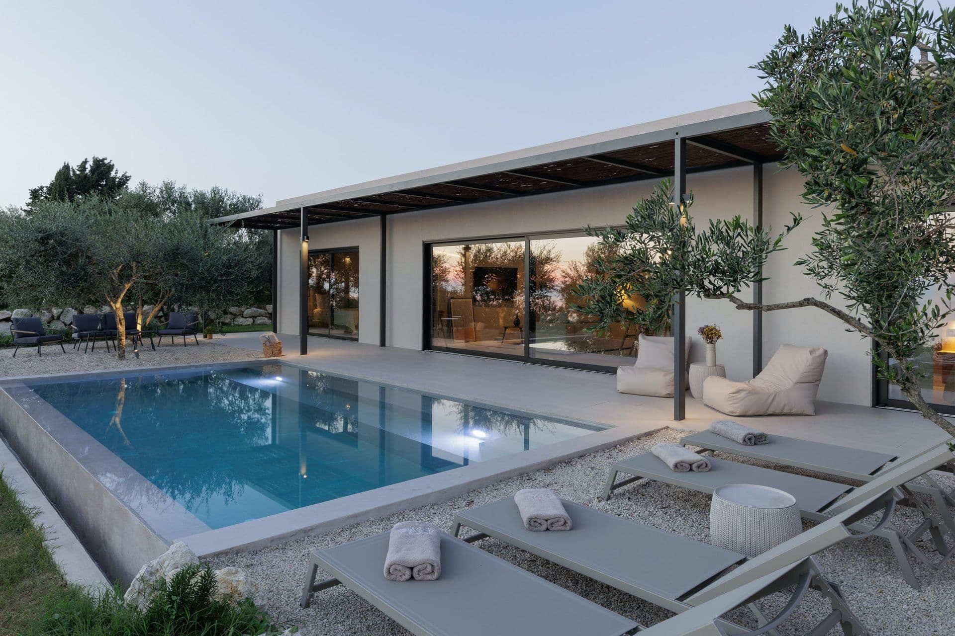 Lit pool and loungers beside the glass villa at dusk at Corfu Dreams, Corfu