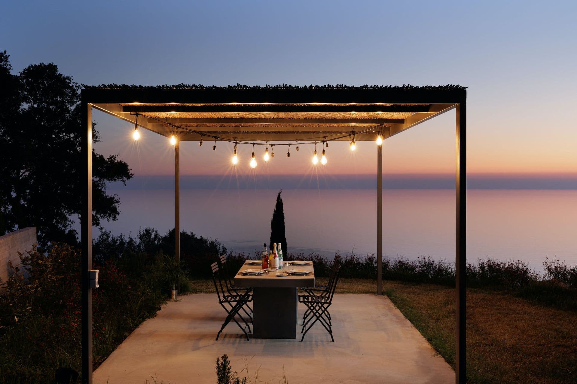 Outdoor dining under a festoon-lit pergola at sunset over the sea at Corfu Dreams, Corfu