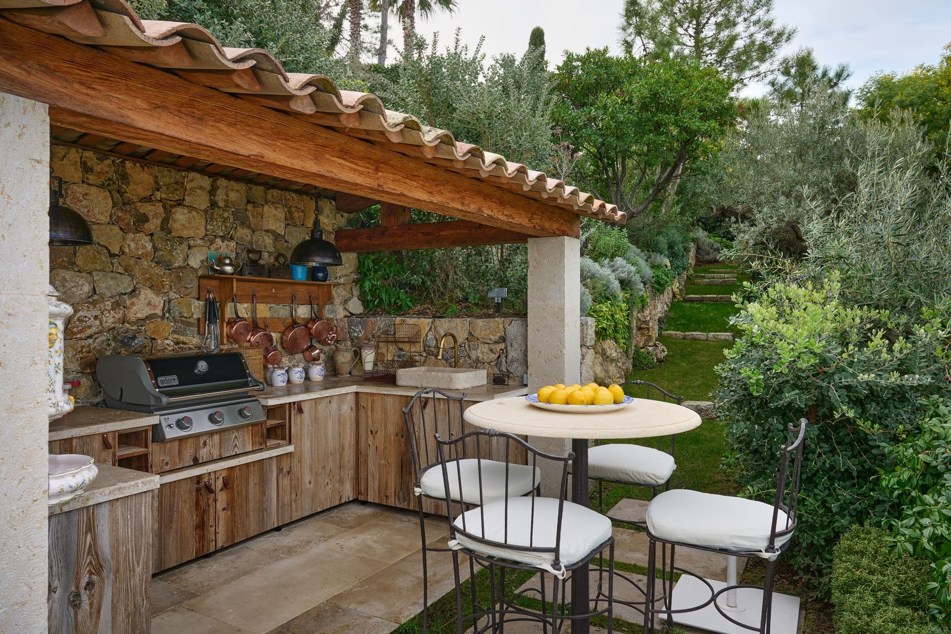 An outdoor kitchen and dining area beneath a tiled pergola surrounded by olive trees