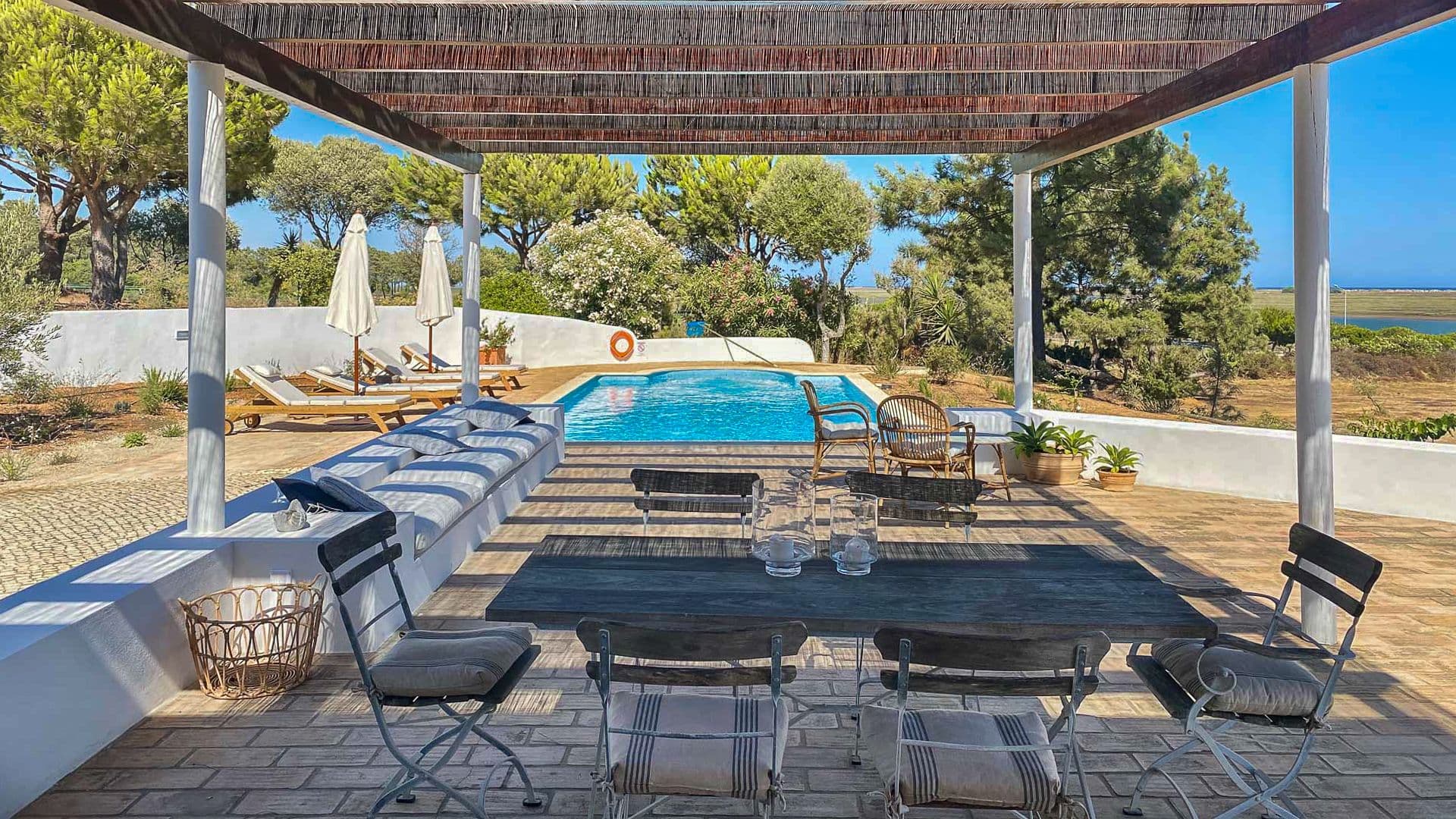 Covered outdoor dining terrace beside the pool, shaded by mature pine trees