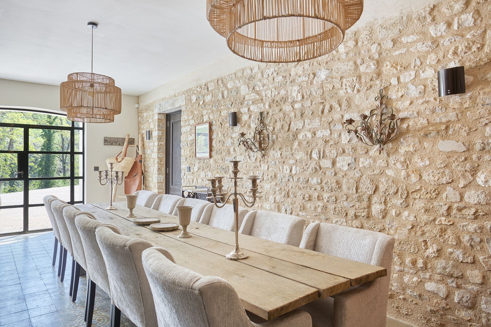 Long banquet dining table for fourteen against an exposed stone wall with rattan pendants at A Glass of Champagne, Provence