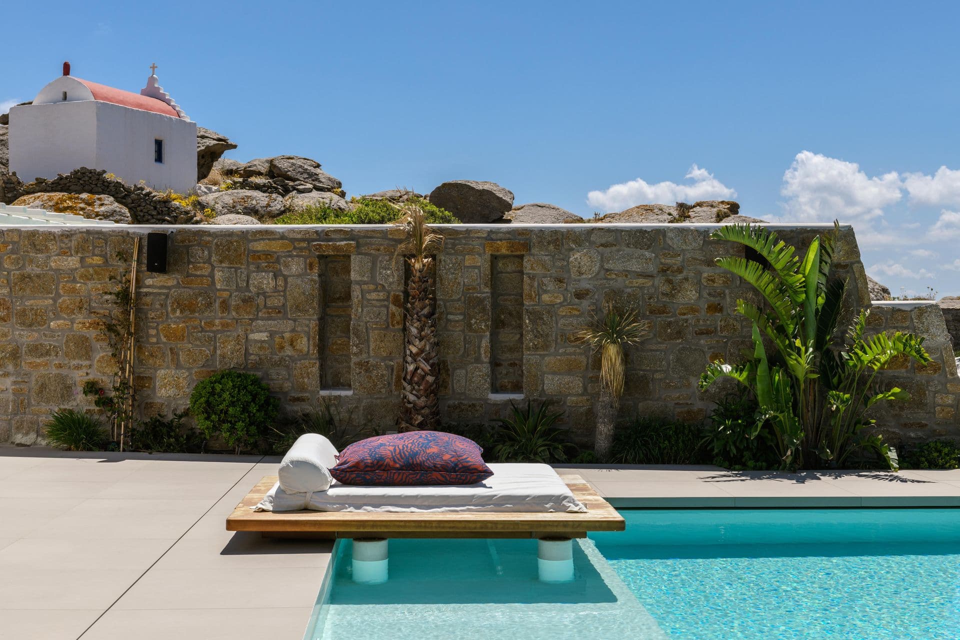 Pool terrace with a stone wall, day bed and a whitewashed chapel behind at Cycladic Sunset Stay, Mykonos