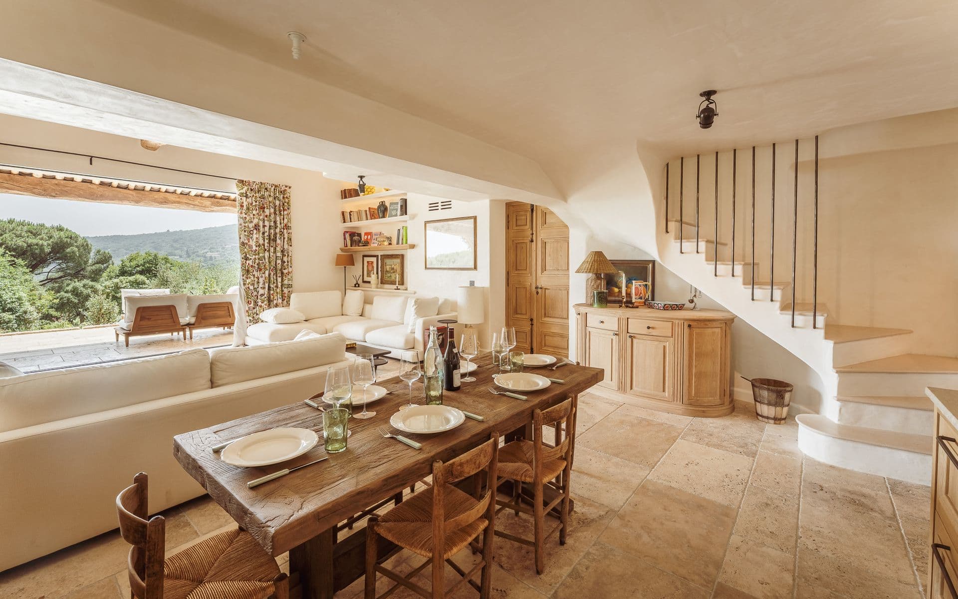 Long wooden dining table set for a group beneath a staircase and beamed ceiling