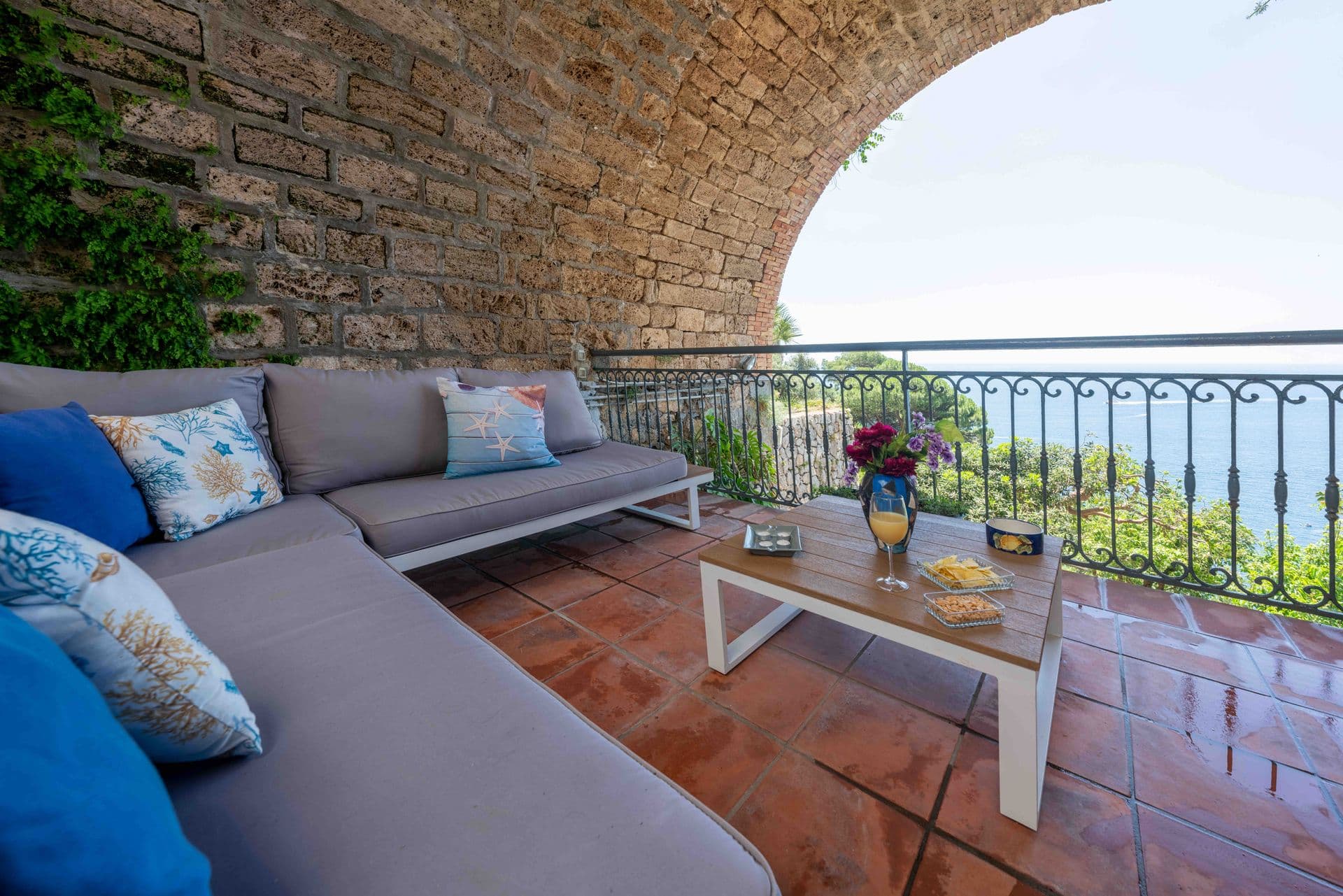 A shaded terrace lounge with grey sofas and cushions overlooking the coast