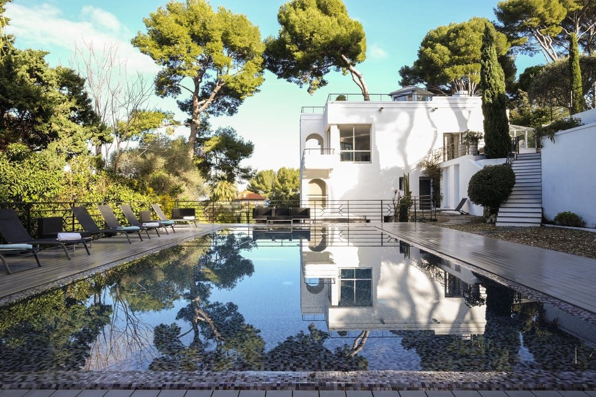 White art-deco villa reflected in a long dark pool surrounded by pine trees