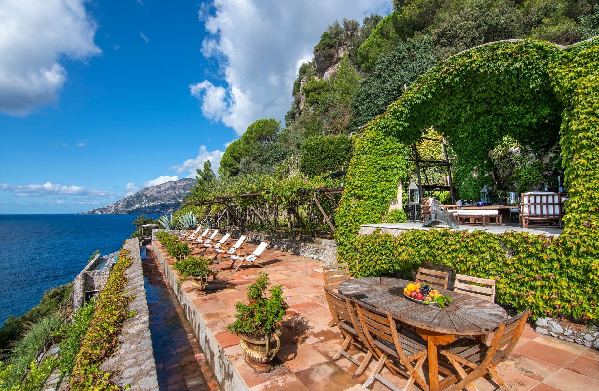 Long stone terrace with dining table and loungers beneath ivy-clad arches
