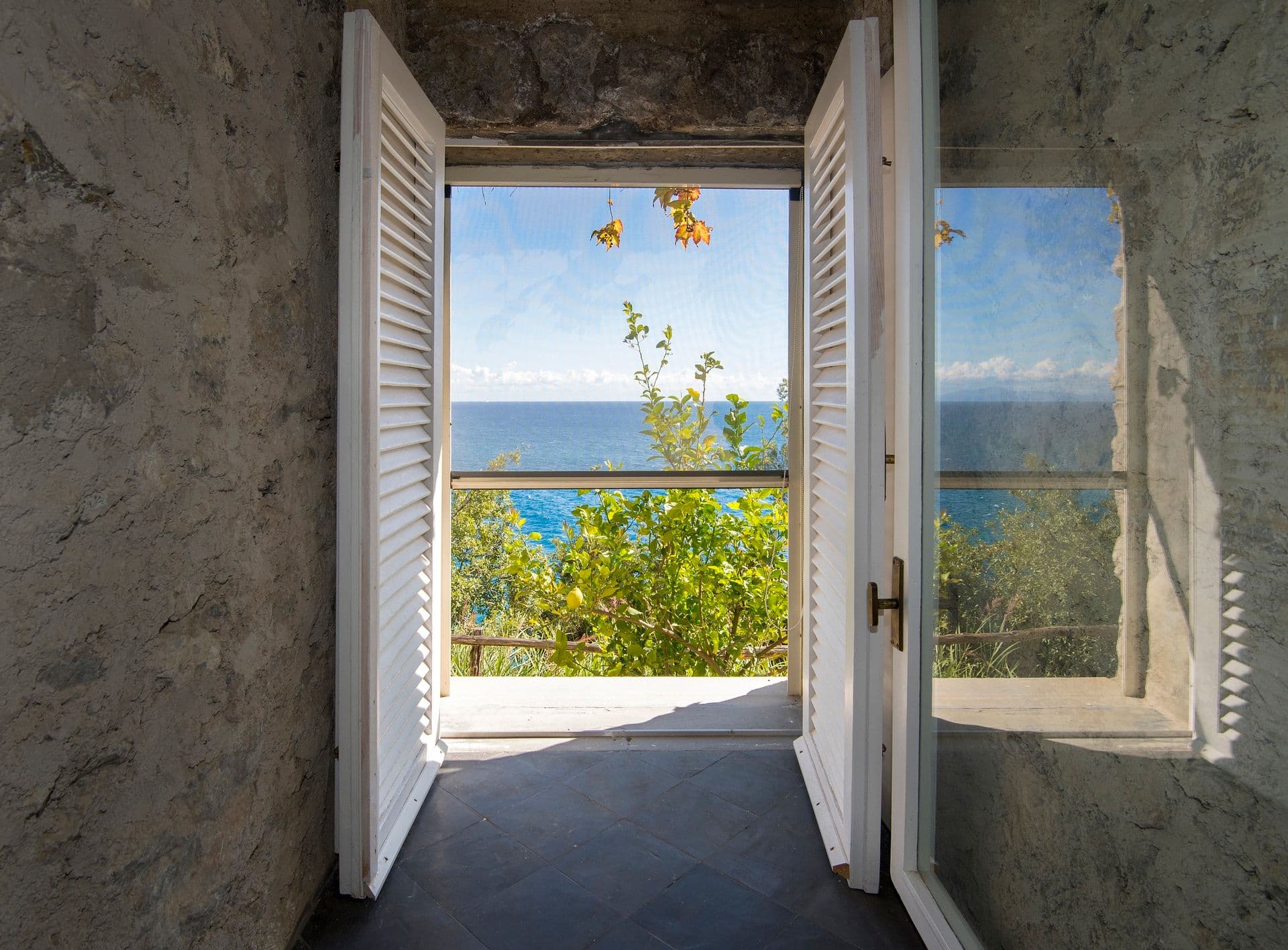 Open shuttered window framing a view of the sea and coastline