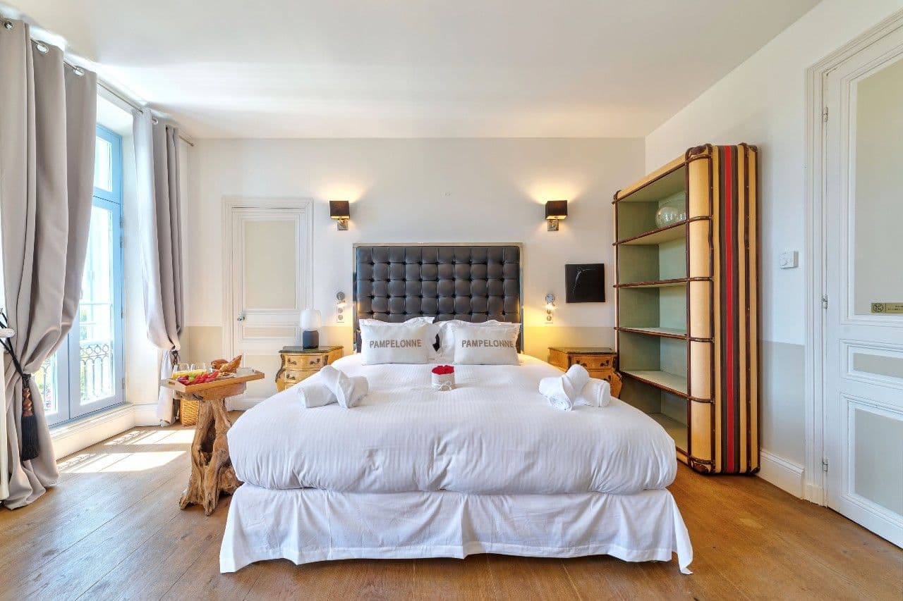 A bright master bedroom with a dark upholstered headboard and tall shuttered windows