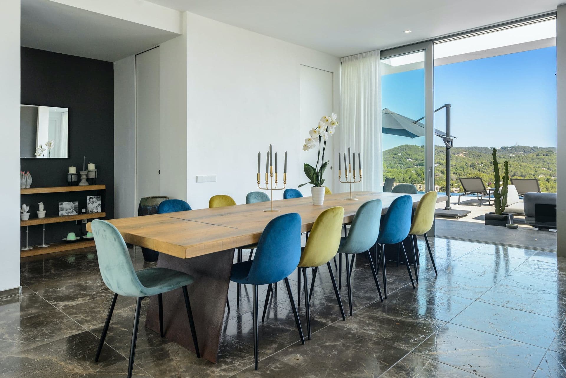 Dining table with mismatched teal and mustard chairs beside floor-to-ceiling windows