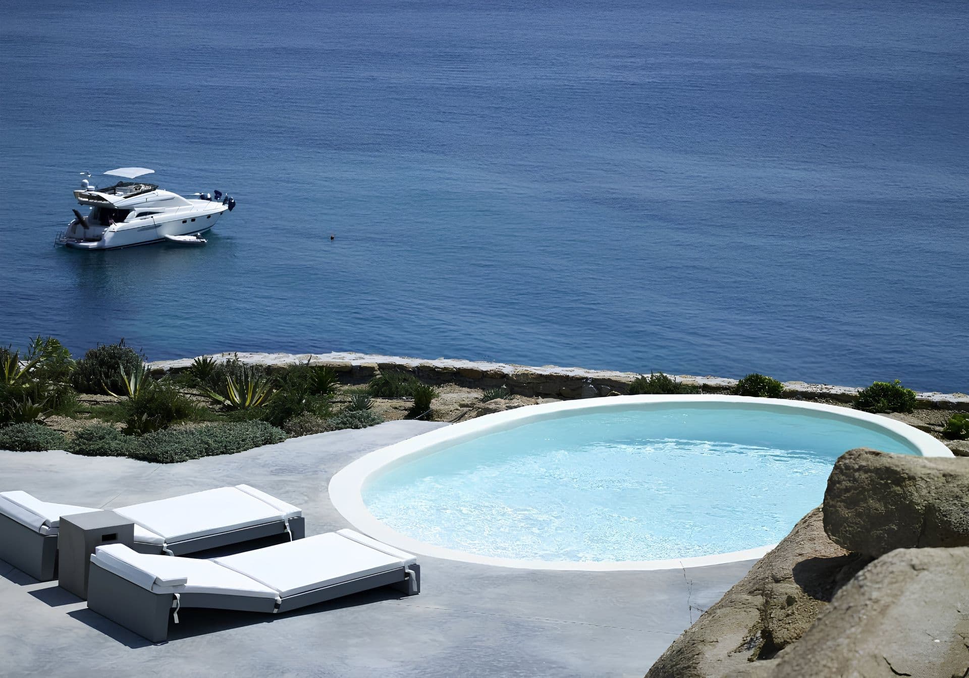 Small round plunge pool on a rocky terrace with a yacht anchored in the bay beyond.