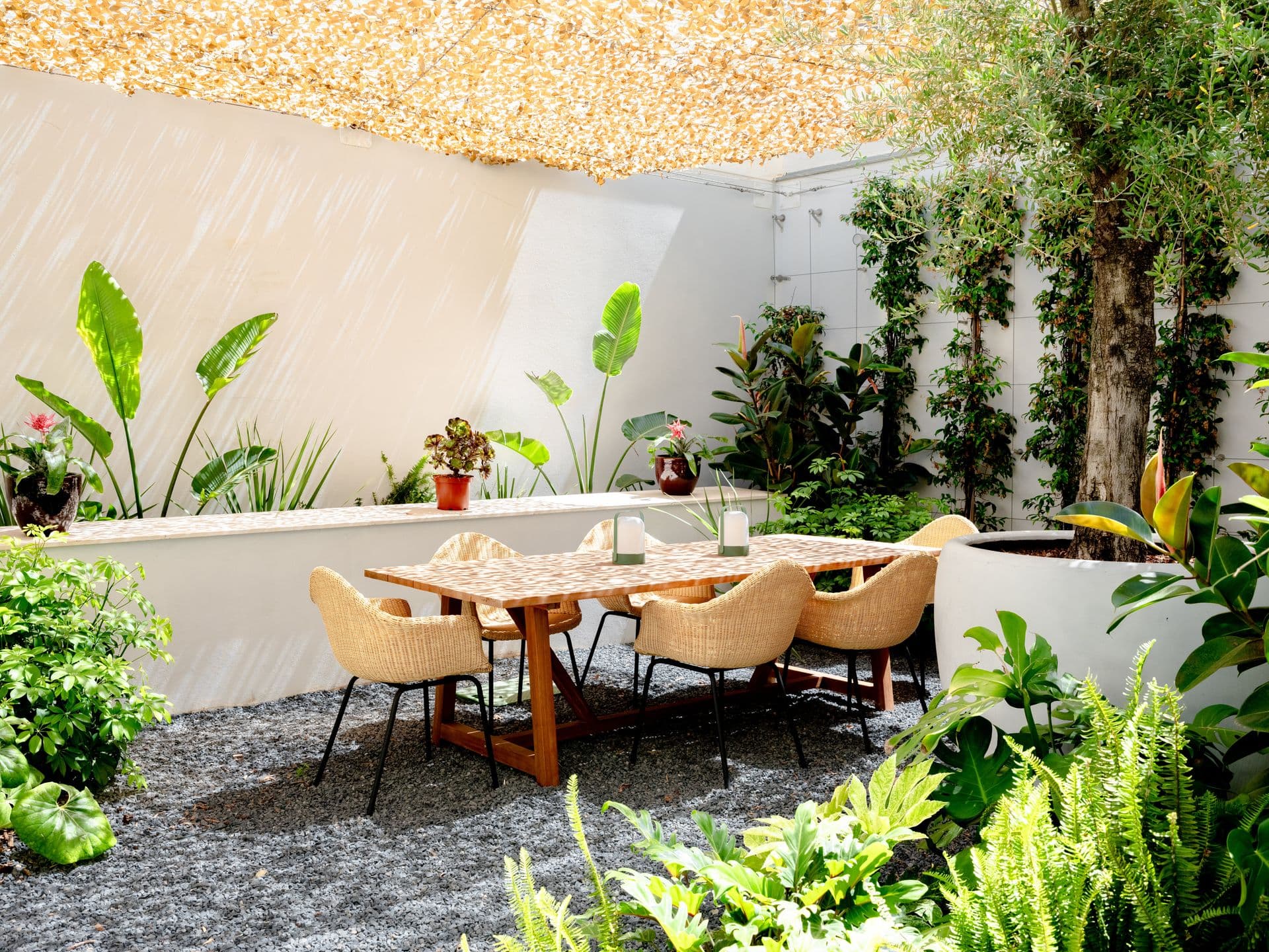 Wooden outdoor dining table set for guests in a gravel courtyard framed by greenery