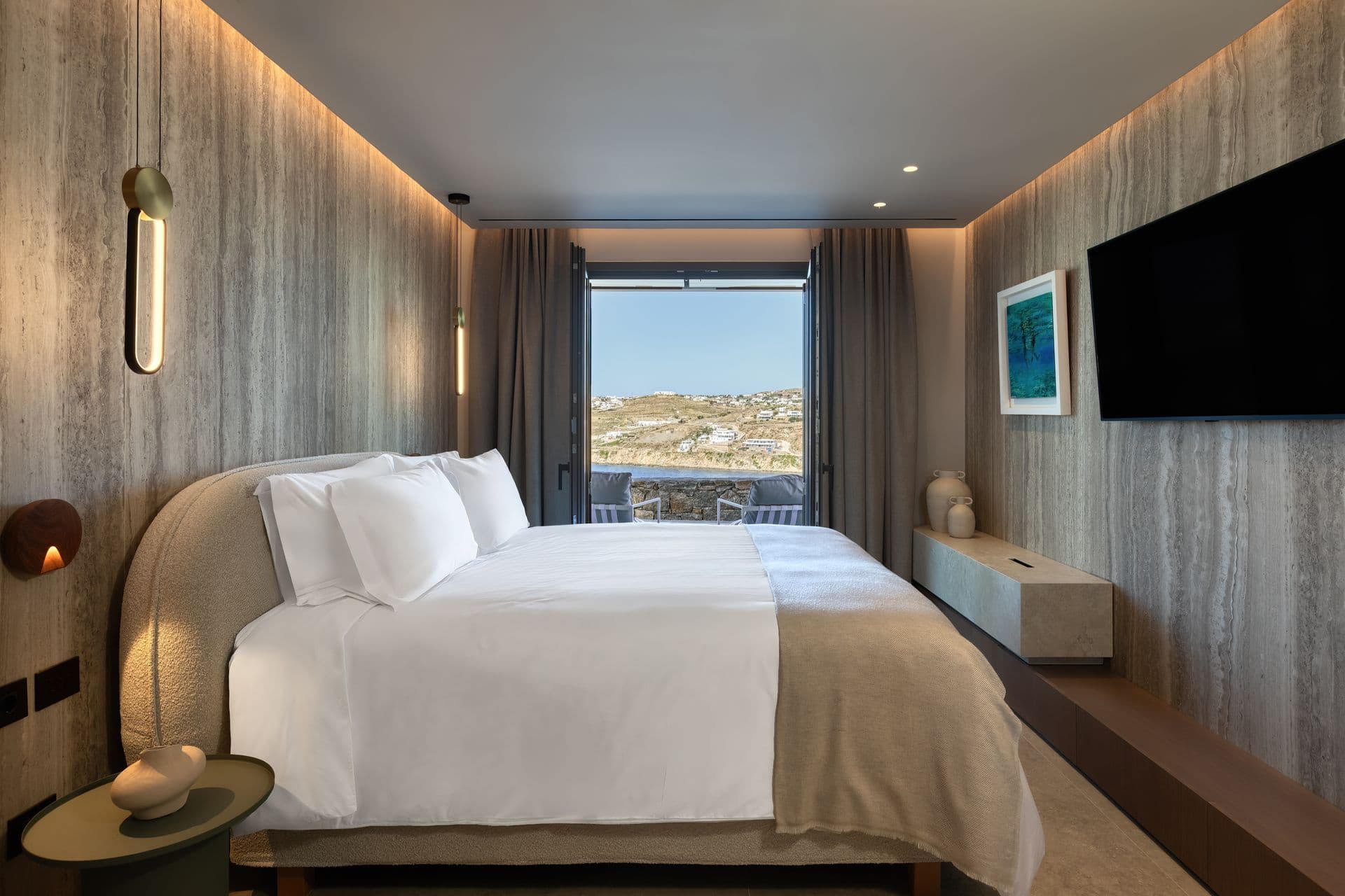Master bedroom with a travertine wall, upholstered bed and sea view at Aegean Aspect, Mykonos