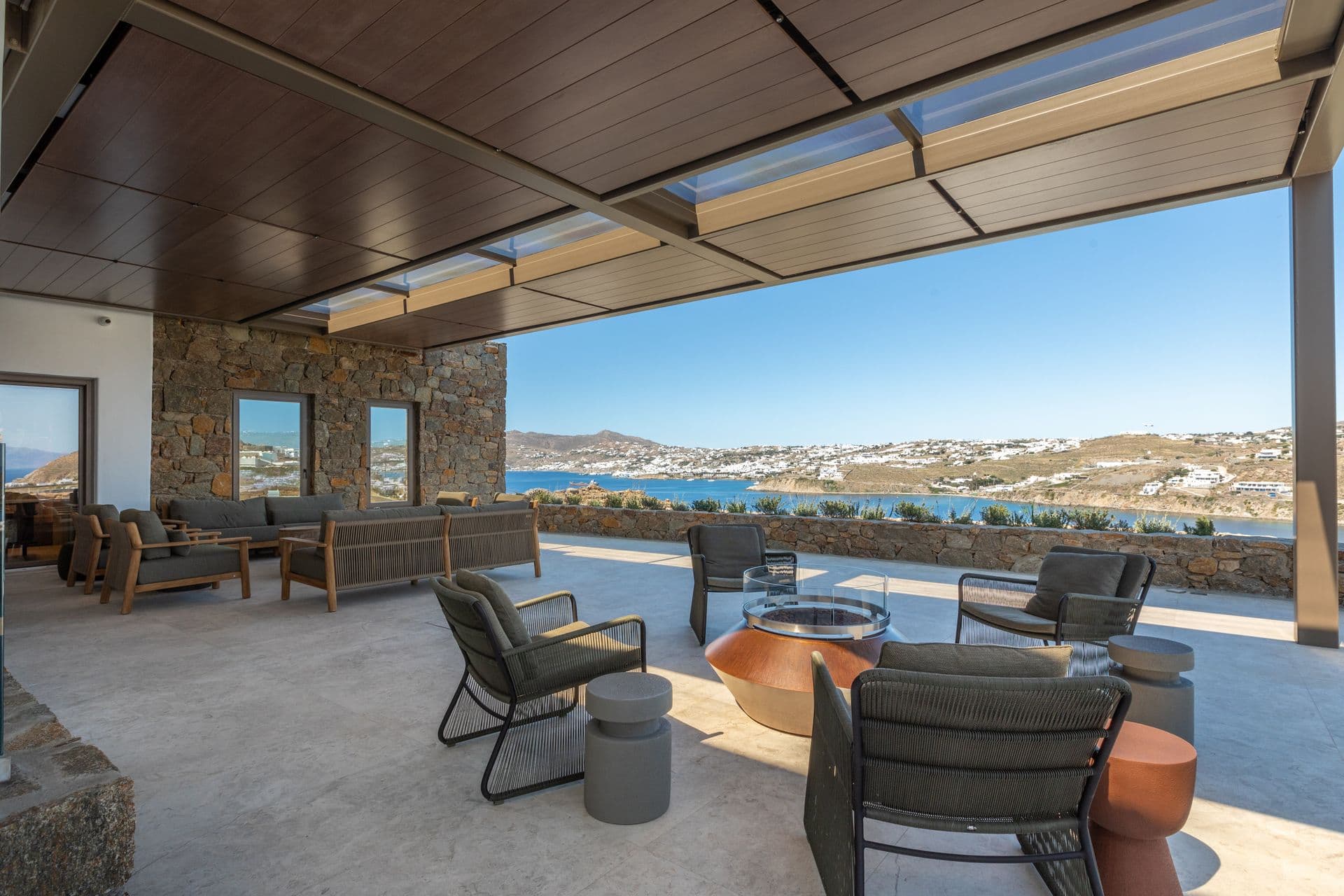 Covered terrace lounge with a firepit and woven chairs above the bay at Aegean Aspect, Mykonos
