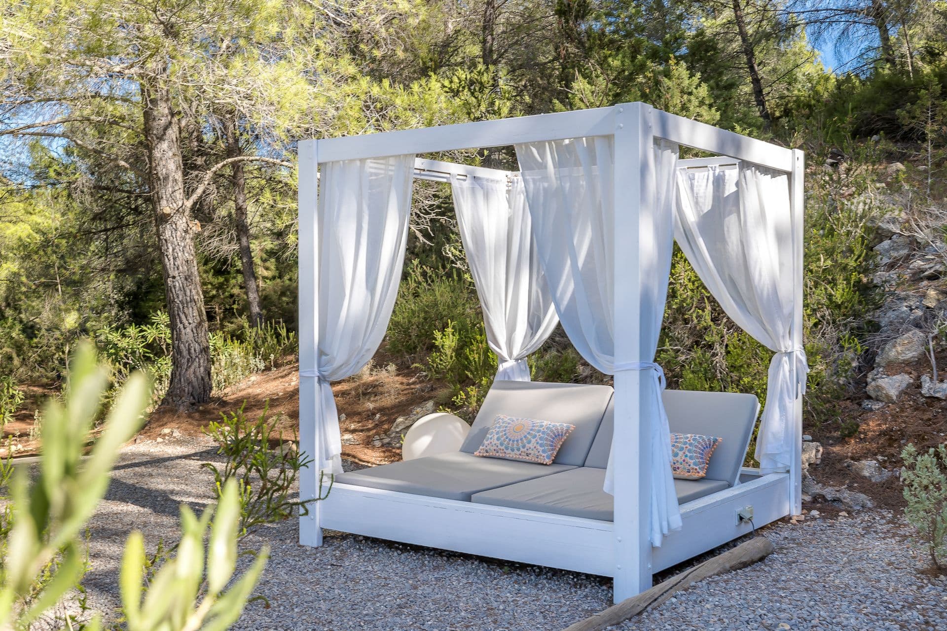 Canopied outdoor daybed set among pine trees in a gravel garden.