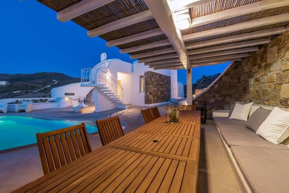 Outdoor dining table under a pergola at dusk with pool and villa beyond