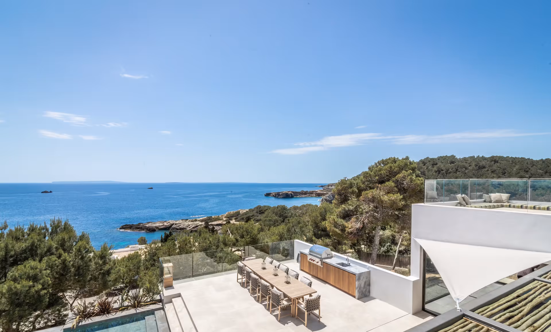 Long outdoor dining table set beneath a pergola with panoramic sea views