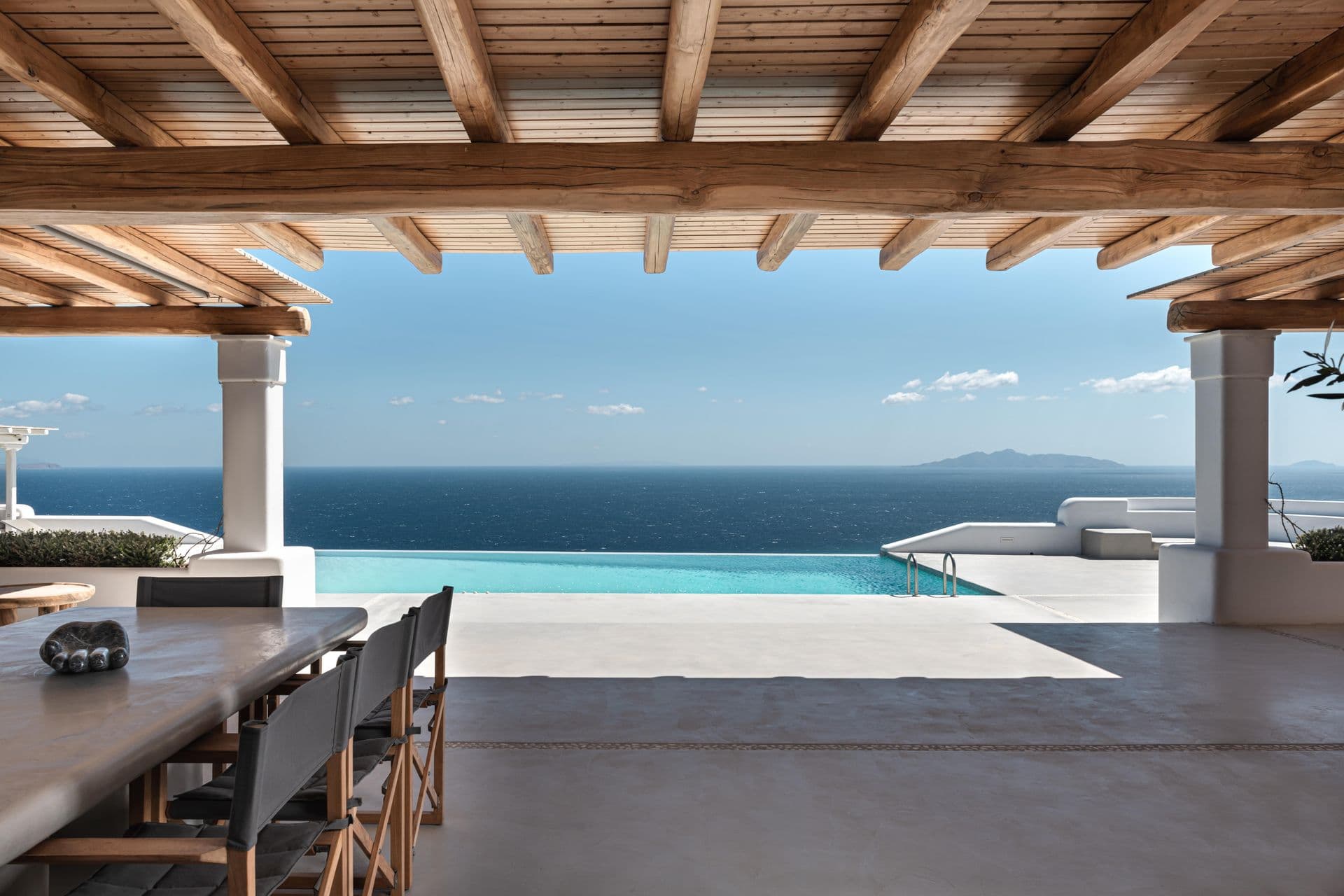 Long outdoor dining table beneath a beamed pergola with uninterrupted Aegean Sea views