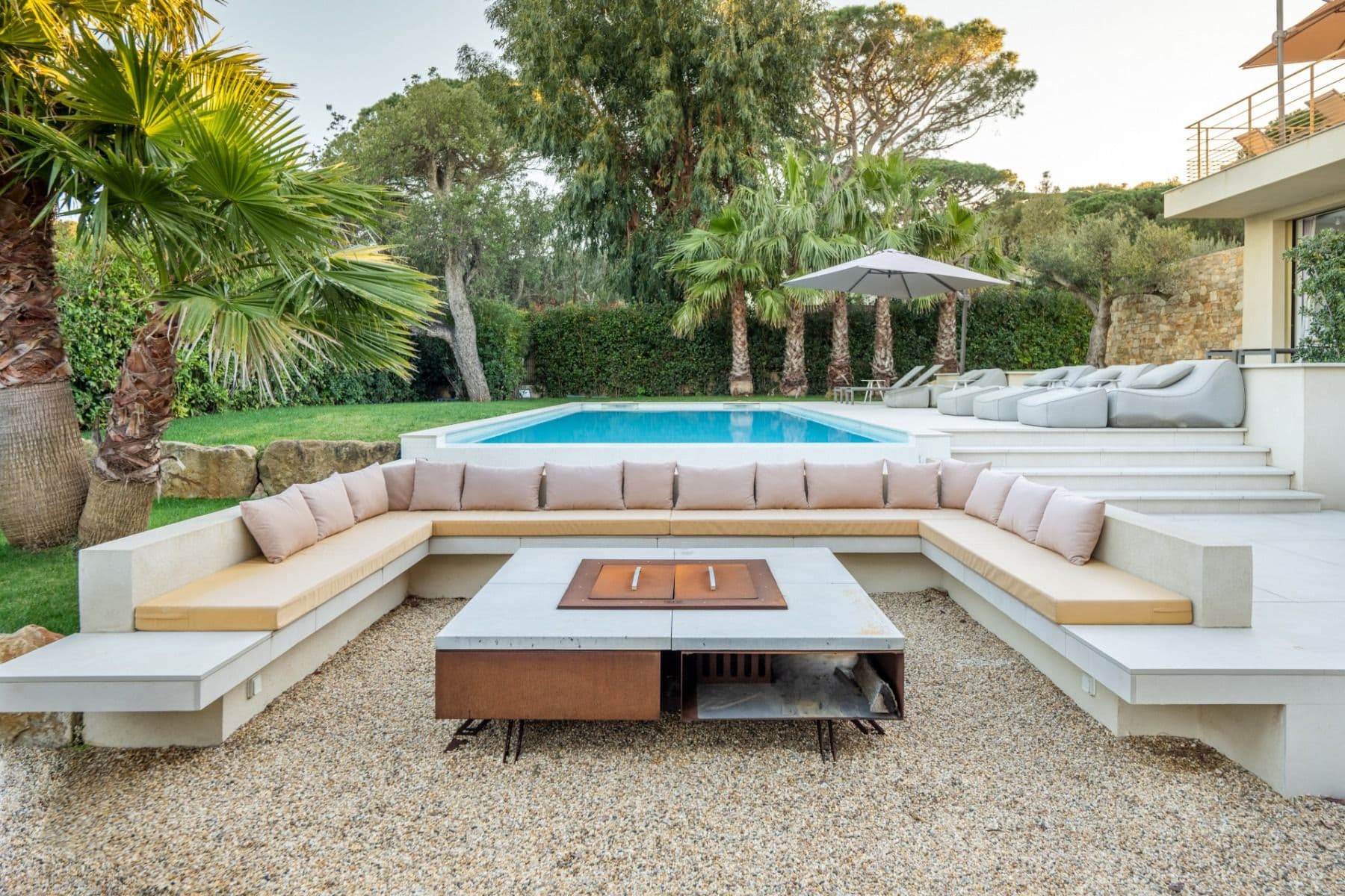 Sunken outdoor lounge seating around a fire pit table beside the pool, framed by palm trees