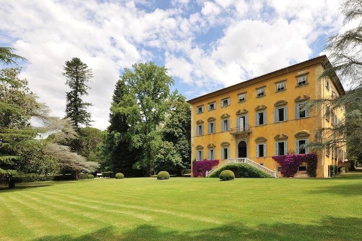 Ochre coloured Tuscan villa facade seen across a manicured lawn framed by mature trees at Midnight Ball
