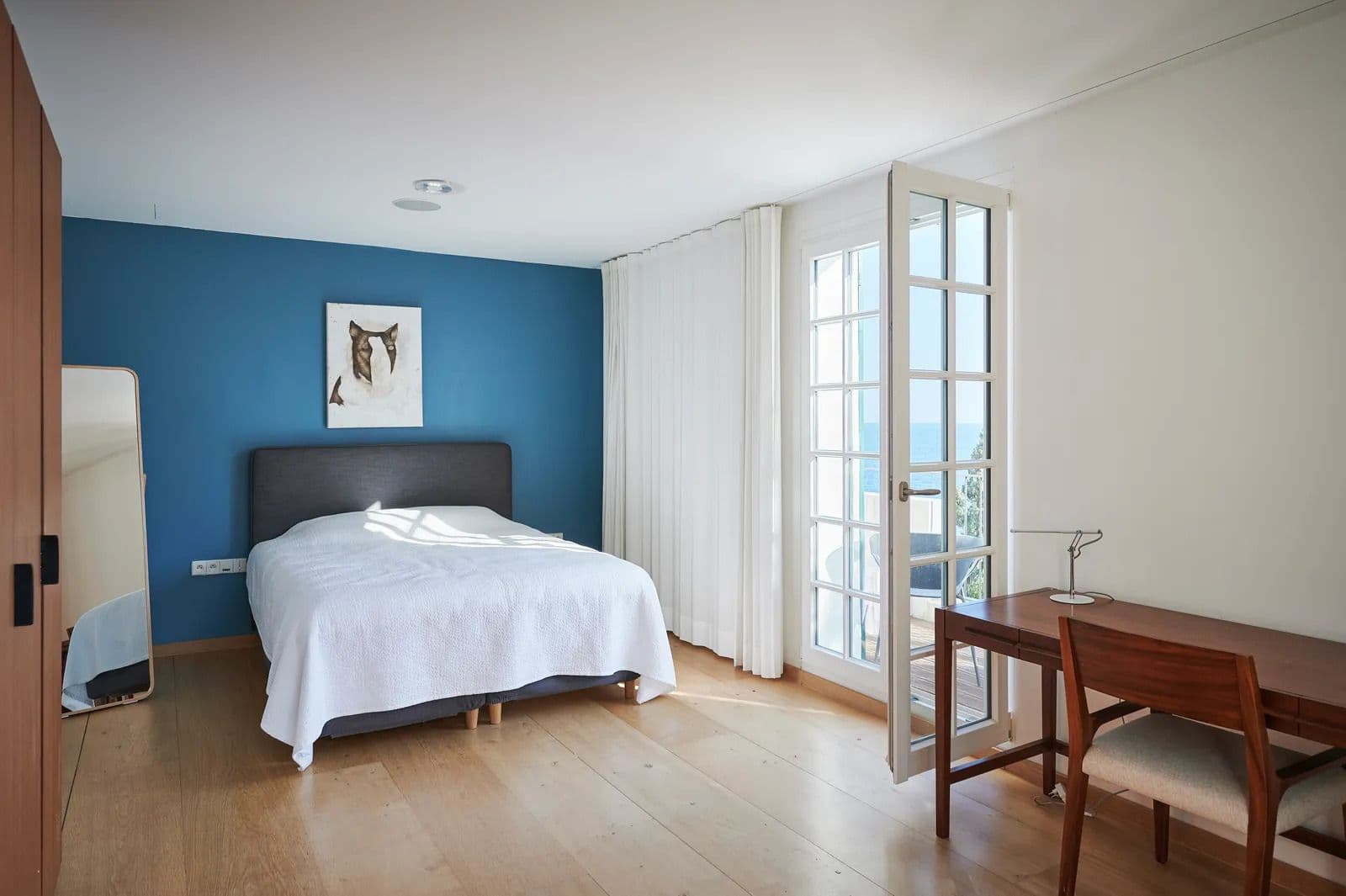 Bedroom with a deep blue accent wall, artwork above the headboard and French doors to a balcony.