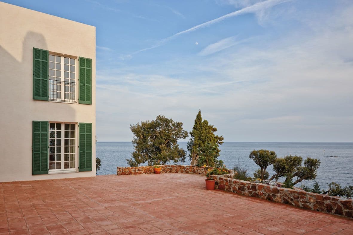 Villa facade with green shutters and a terracotta tiled terrace overlooking the sea.