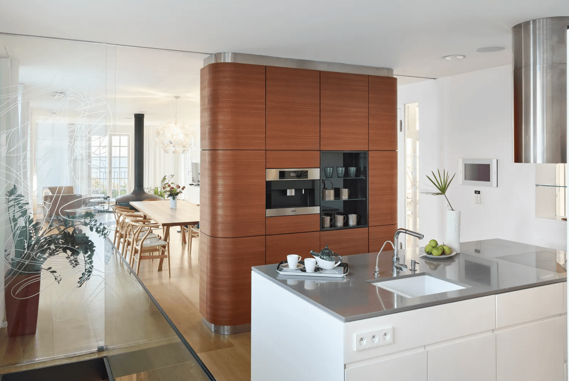 Open kitchen with a curved, wood clad cabinetry wall, a built in coffee machine and a stainless steel island.