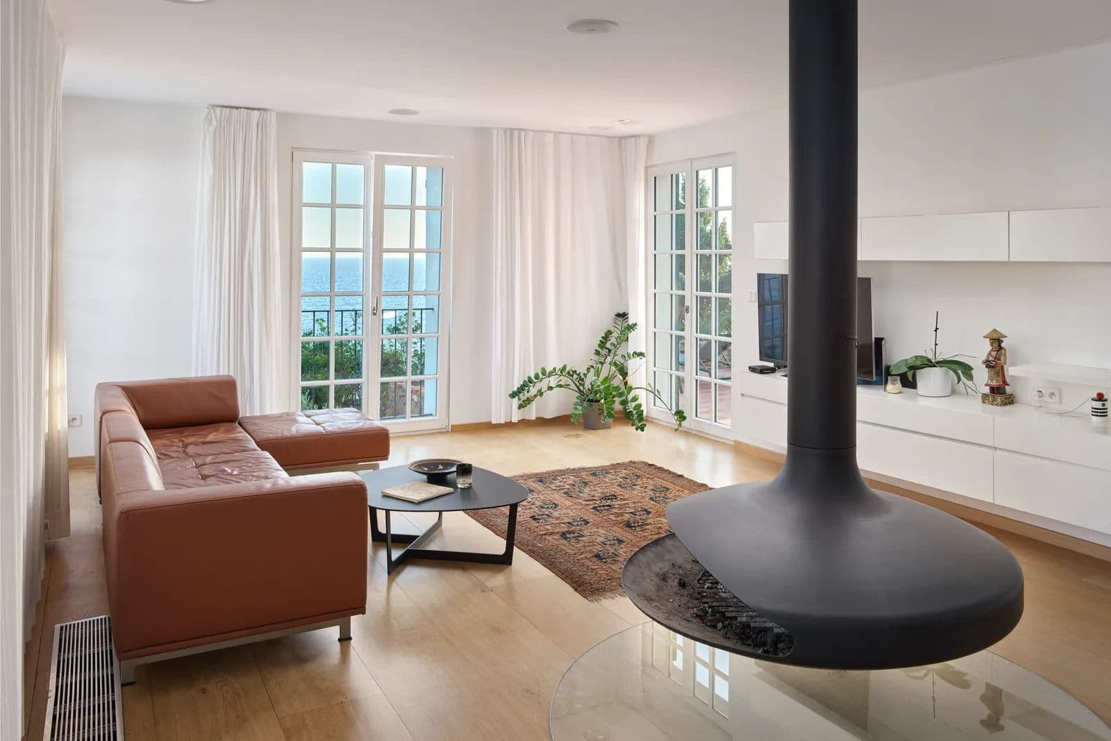 Living room with a tan leather sofa, rug and a suspended black fireplace beside glass doors to the garden.