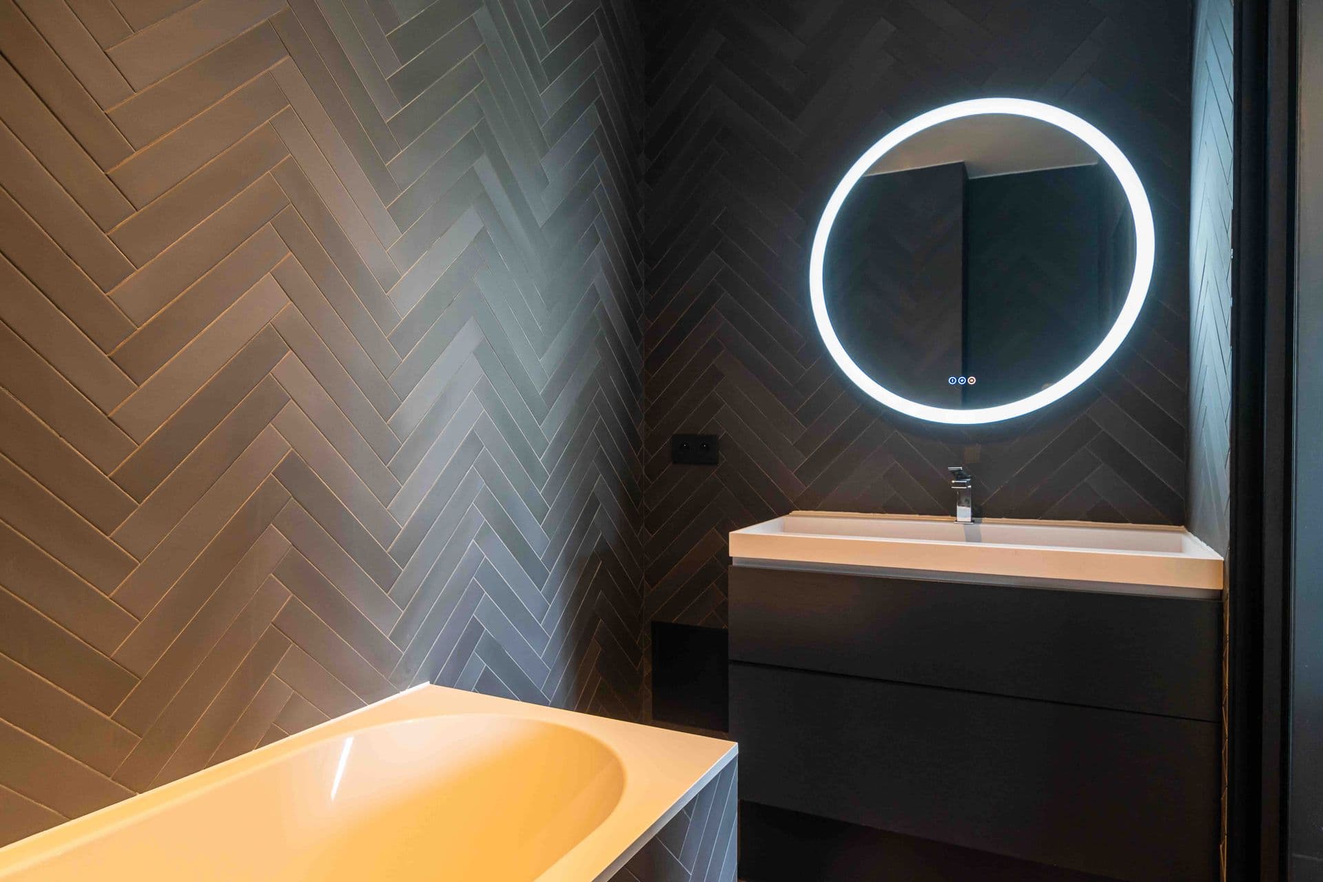 A bathroom with herringbone tiling and a round backlit mirror above the vanity