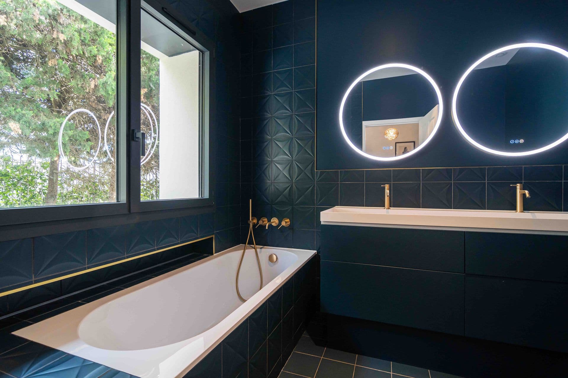 A navy tiled bathroom with a double vanity and two round illuminated mirrors