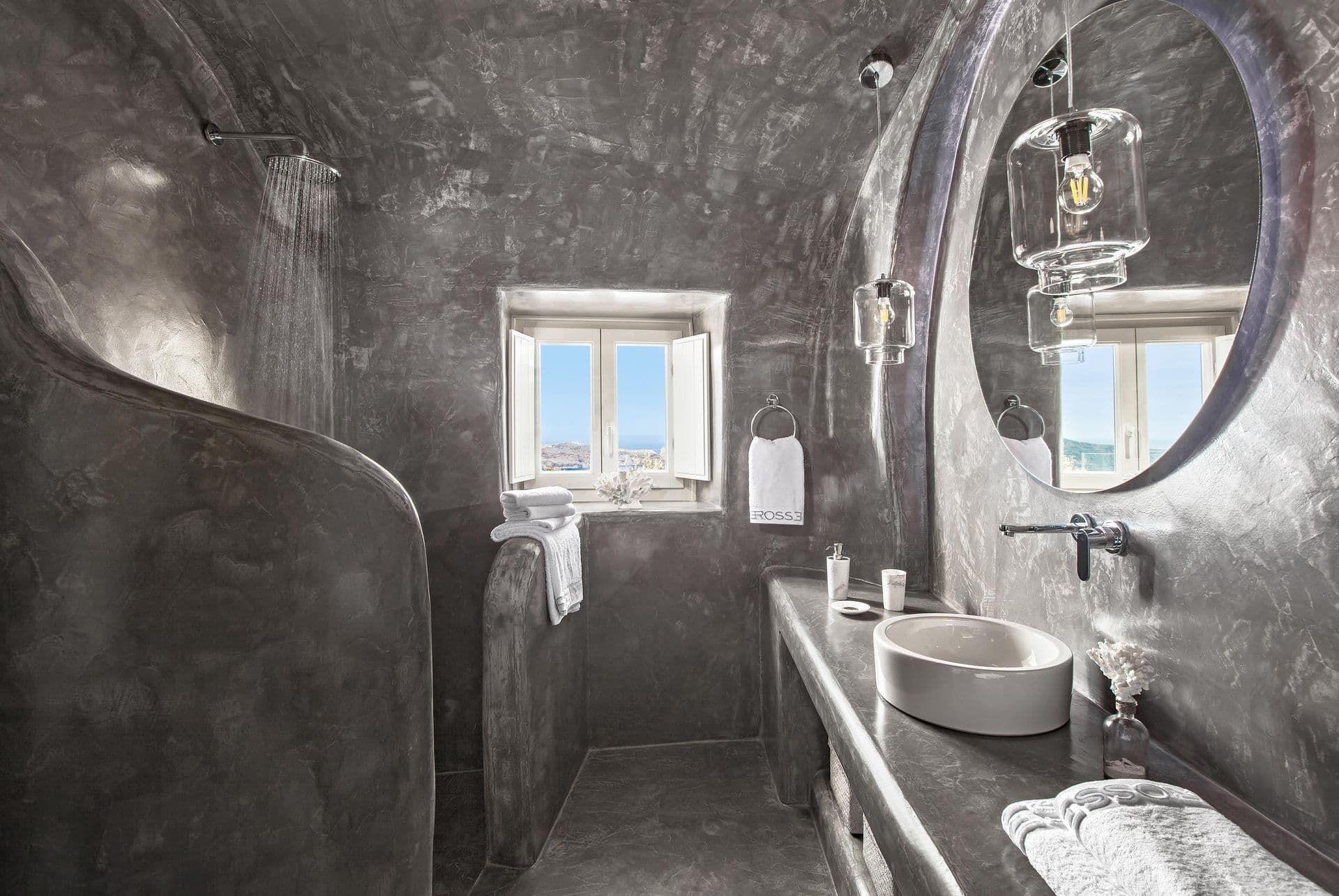 Grey microcement bathroom with a round mirror and a sea-view window at Cave Chic, Imerovigli, Santorini
