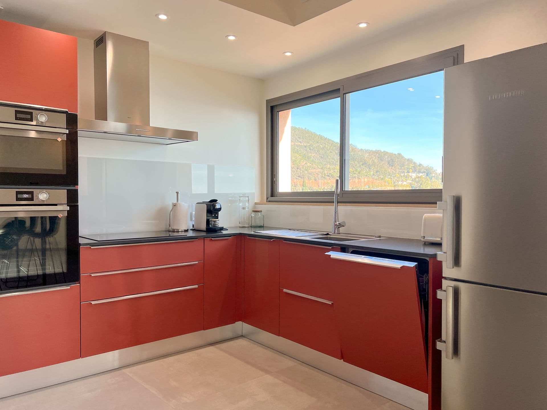 Fitted kitchen with red cabinetry and integrated appliances beside a sea-view window