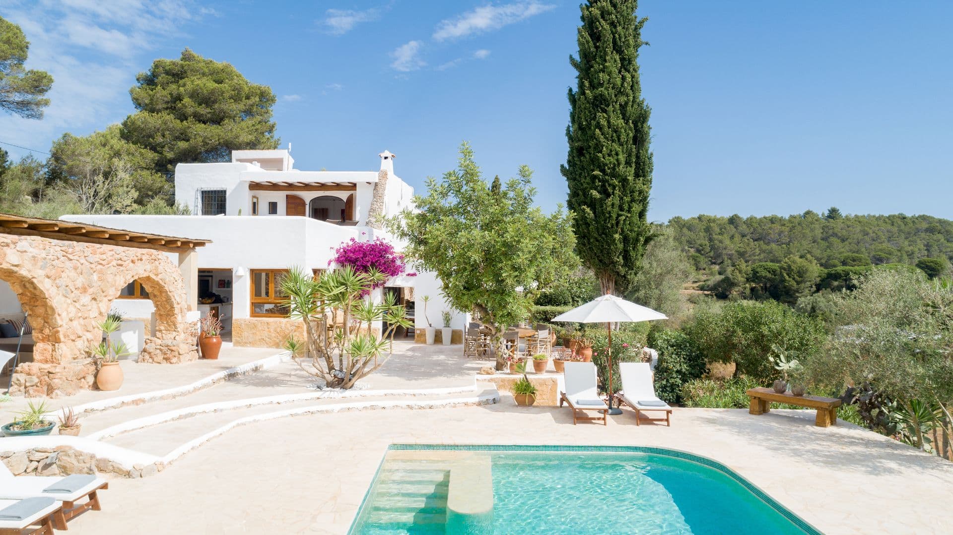 Whitewashed Ibizan finca with swimming pool, terrace and surrounding pine trees under blue sky